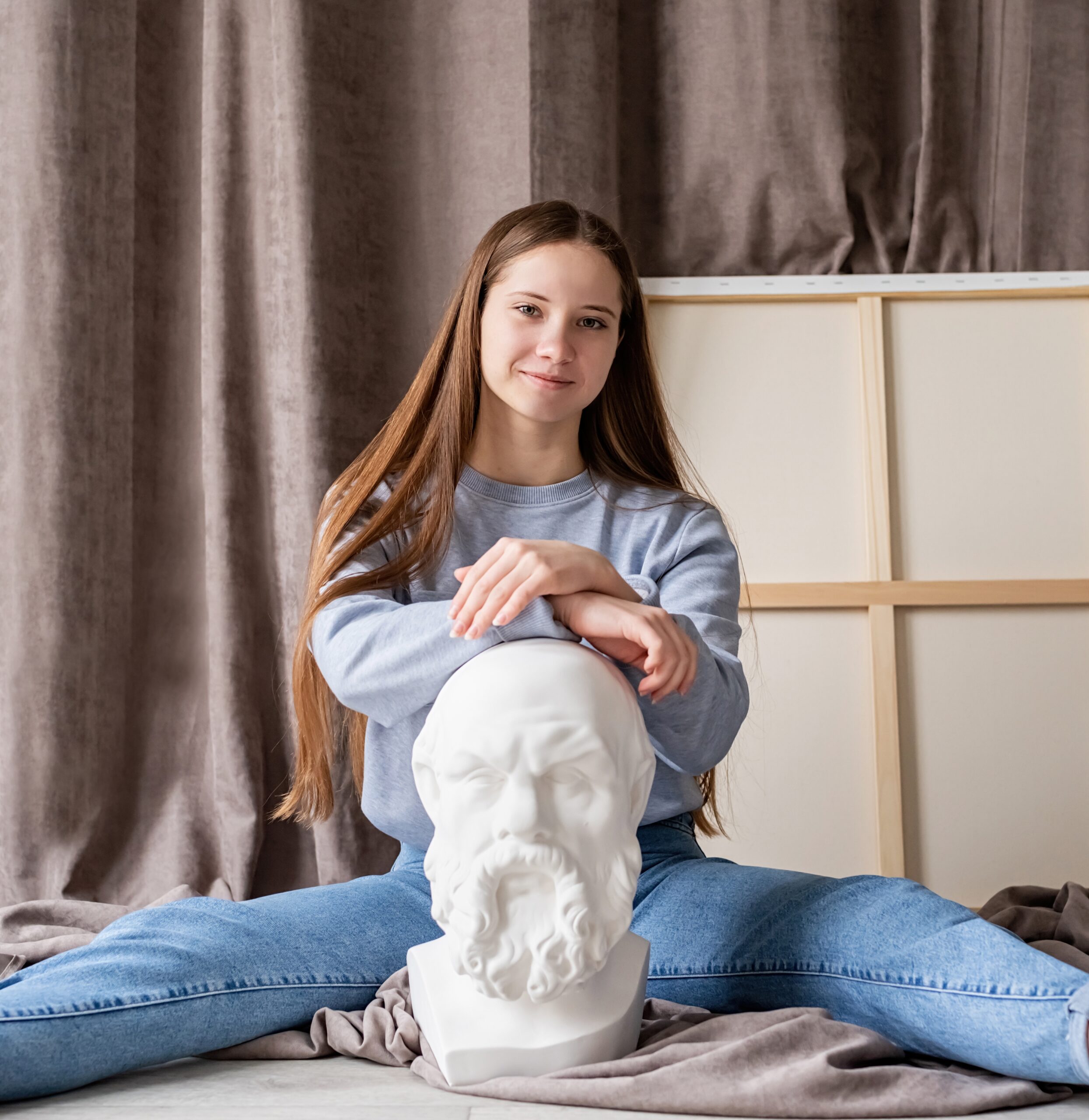 Young smiling scholar sitting in a studio with the gypsum sculpture of Socrates head and canvas